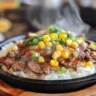 A sizzling hot plate of homemade beef pepper rice with thinly sliced beef, white rice, corn, green onions, and black pepper, served on a cast iron skillet with steam rising.