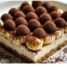 "A slice of Banana Pudding Tiramisu on a white plate, showing layers of espresso-soaked ladyfingers, creamy mascarpone, fresh banana slices, and vanilla wafers, topped with dark chocolate shavings."