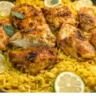 A close-up of turmeric chicken and rice garnished with fresh herbs and lime slices. The golden yellow rice contrasts with the crispy, spiced chicken pieces.